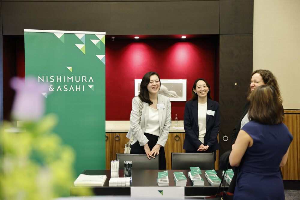 Vier Frauen stehen im Gespräch an einem Messestand, Tisch mit Broschüren und Stiften, hinter ihnen ein grüner Banner mit der Aufschrift Nishimura & Asahi.