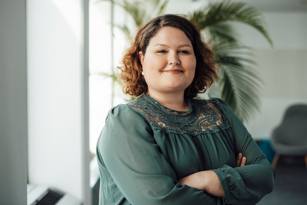 A portrait photograph of a young woman wearing an olive green blouse. She has her arms crossed.