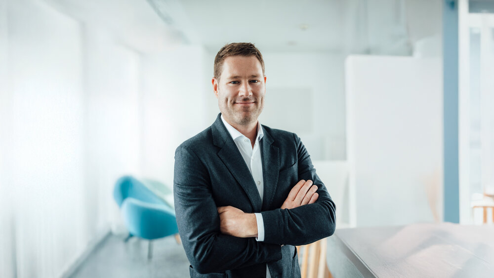 A portrait photograph of a smartly dressed man with styled light brown hair and his arms crossed in an office space.