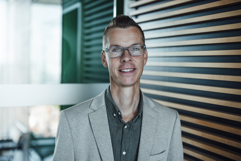 A portrait photograph of a man with short brown styled hair standing in front of a glass partition. He is wearing glasses.