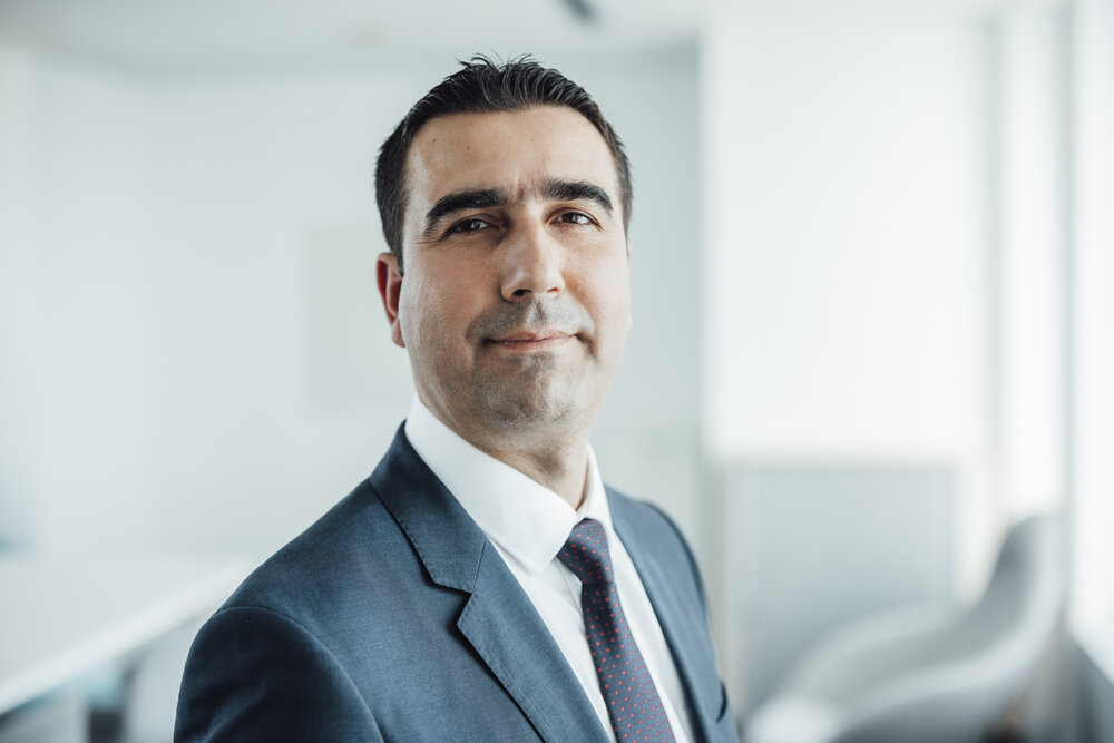 A portrait photograph of a smartly dressed man standing sideways in an office.