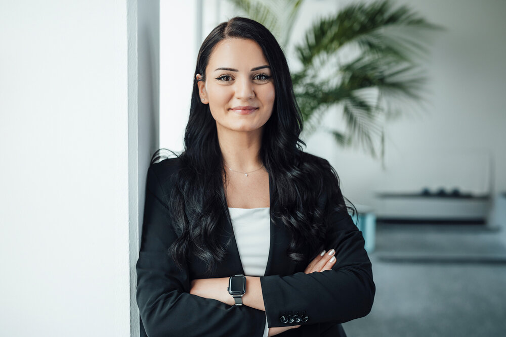 A portrait photograph of a woman standing sideways against an office wall. She is wearing a white top and a black blazer.