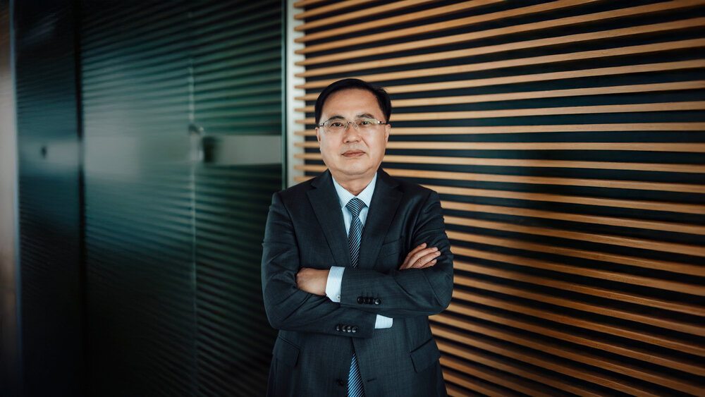 A portrait photograph of a smartly dressed man standing in front of an office wall.