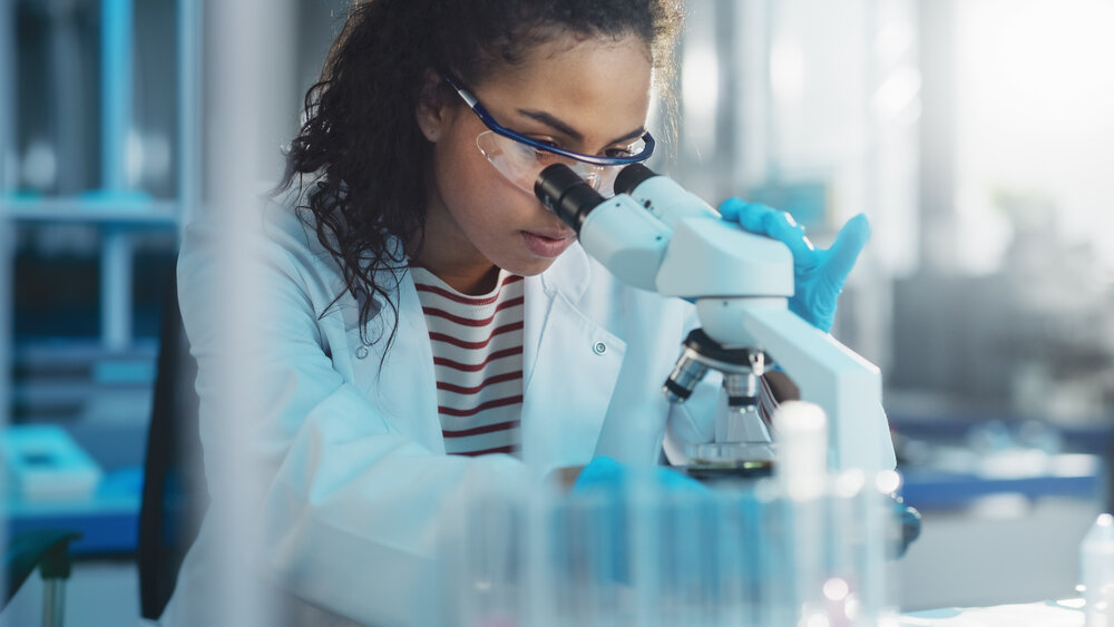 A woman wearing safety goggles looks through a microscope in a laboratory.