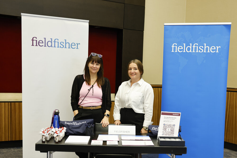 Zwei Frauen stehen hinter einem Messestand mit Broschüren und Giveaways, im Hintergrund Banner mit der Aufschrift fieldfisher.