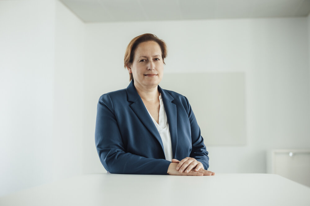 A portrait photo of a smartly dressed woman in a velvet blue blazer. She is sitting behind an office desk with her hands resting on it.