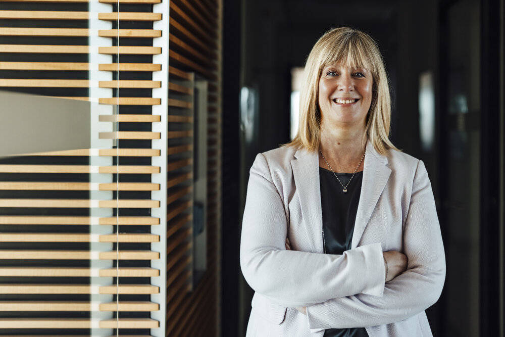 A portrait photograph of a smartly dressed woman. She has her arms crossed and is wearing a light grey blazer and a necklace.