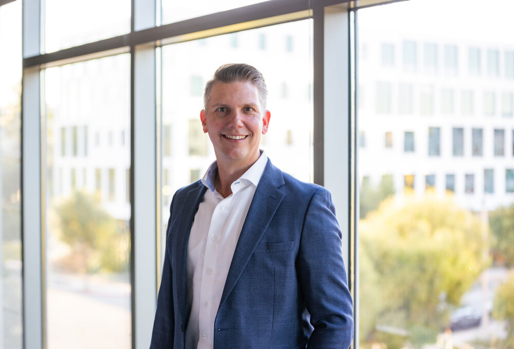 A portrait photograph of a smartly dressed man with styled hair standing in front of several windows.