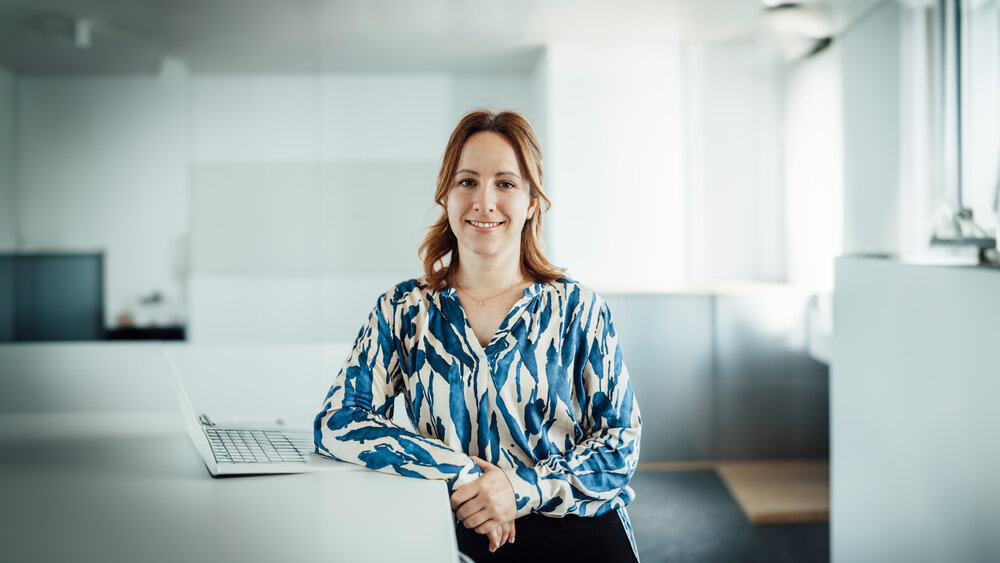 A portrait photo of a young woman. She is wearing a patterned blue and white blouse. She is standing at an office desk with a laptop on it.