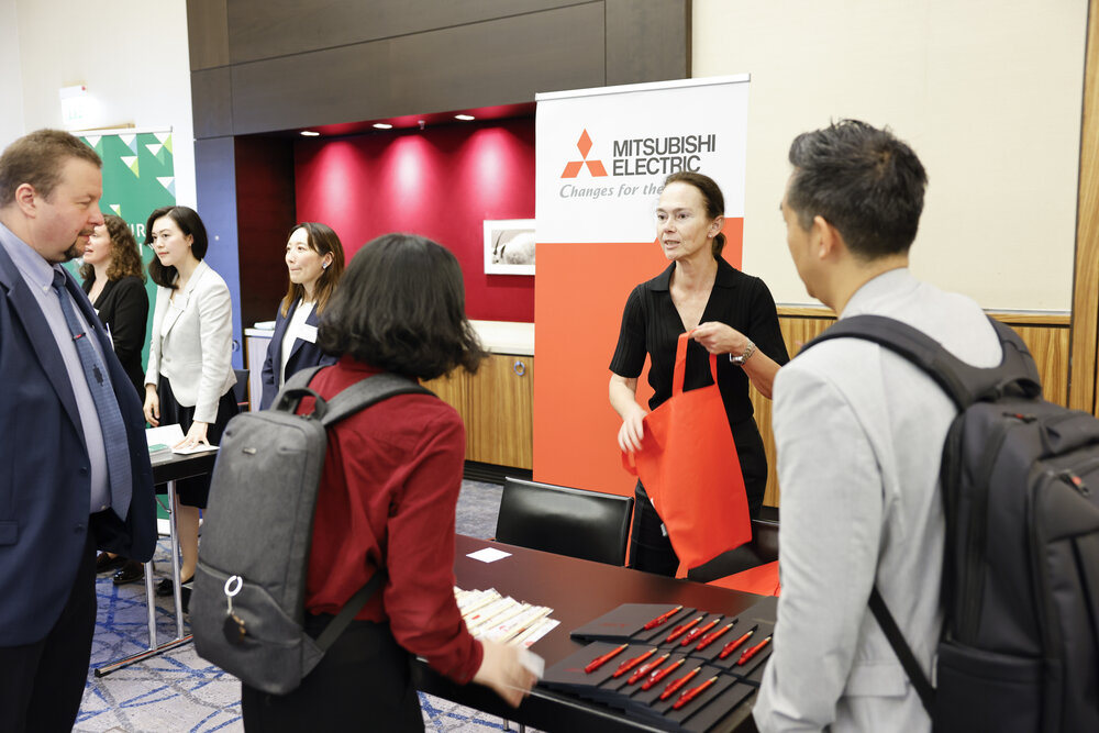 Mehrere Personen an einem Infostand, eine Frau hält eine rote Tasche, im Hintergrund ein Banner von Mitsubishi Electric.